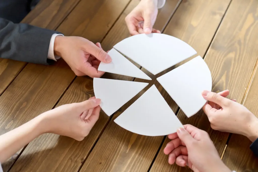 Imagem mostrando mãos de pessoas segurando peças de um gráfico de pizza em uma mesa de madeira, representando colaboração e trabalho em equipe.