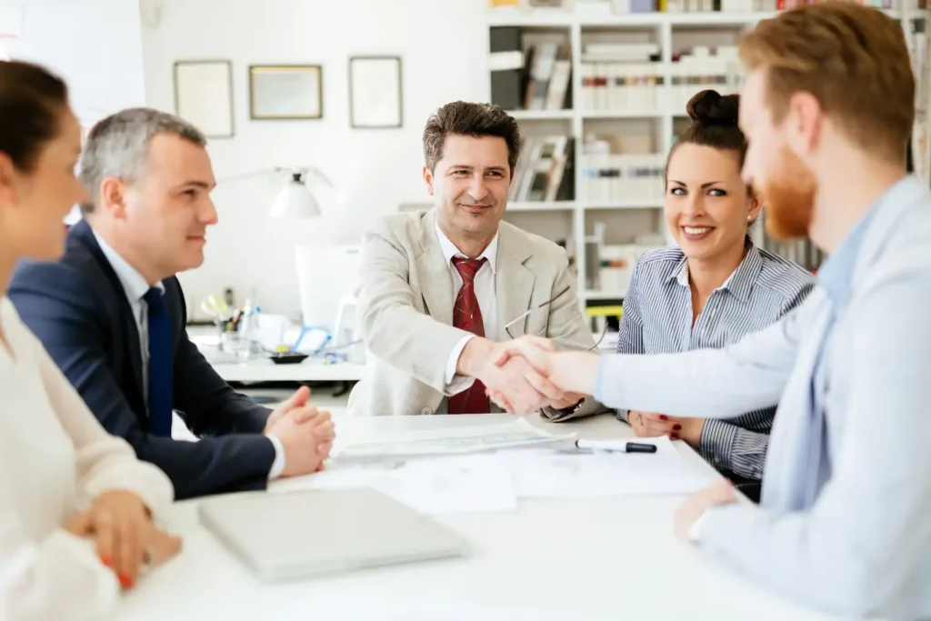 Imagem de uma reunião de negócios com cinco profissionais, onde um homem está apertando a mão de um colega. O ambiente é moderno e colaborativo, refletindo um momento de acordo.