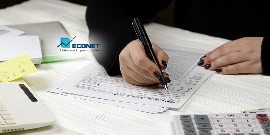 Pessoa assinando documentos importantes na mesa de trabalho, com calculadora, pasta de arquivos e anotações ao lado. Ambiente de escritório organizado.